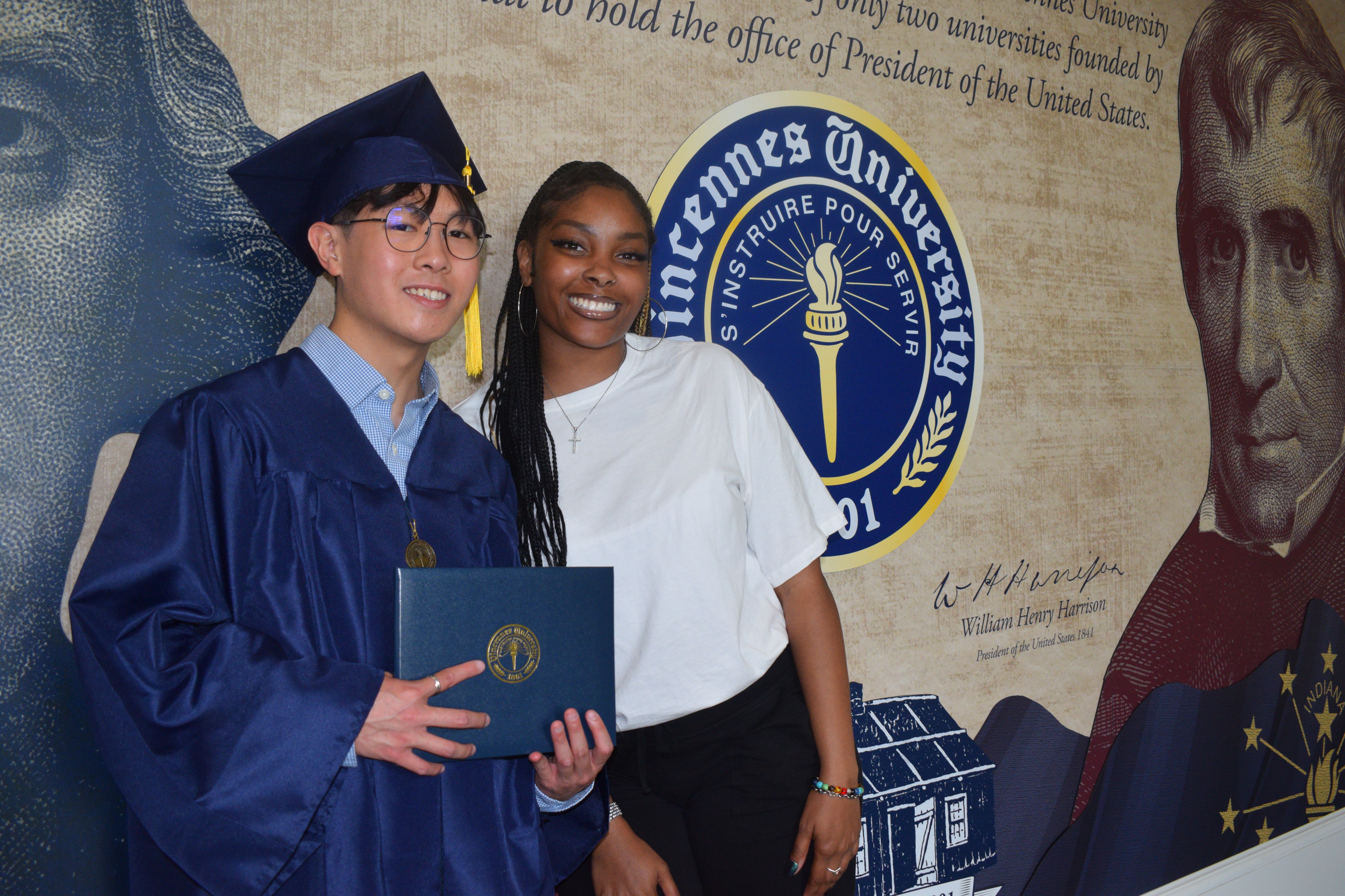 Samuel Lo wears a graduation cap and gown while holding a VU degree cover. A female student stands next to him. The students stand in front of a VU mural featuring Thomas Jefferson, William Henry Harrison, and the VU Seal plus Indiana and American flags.