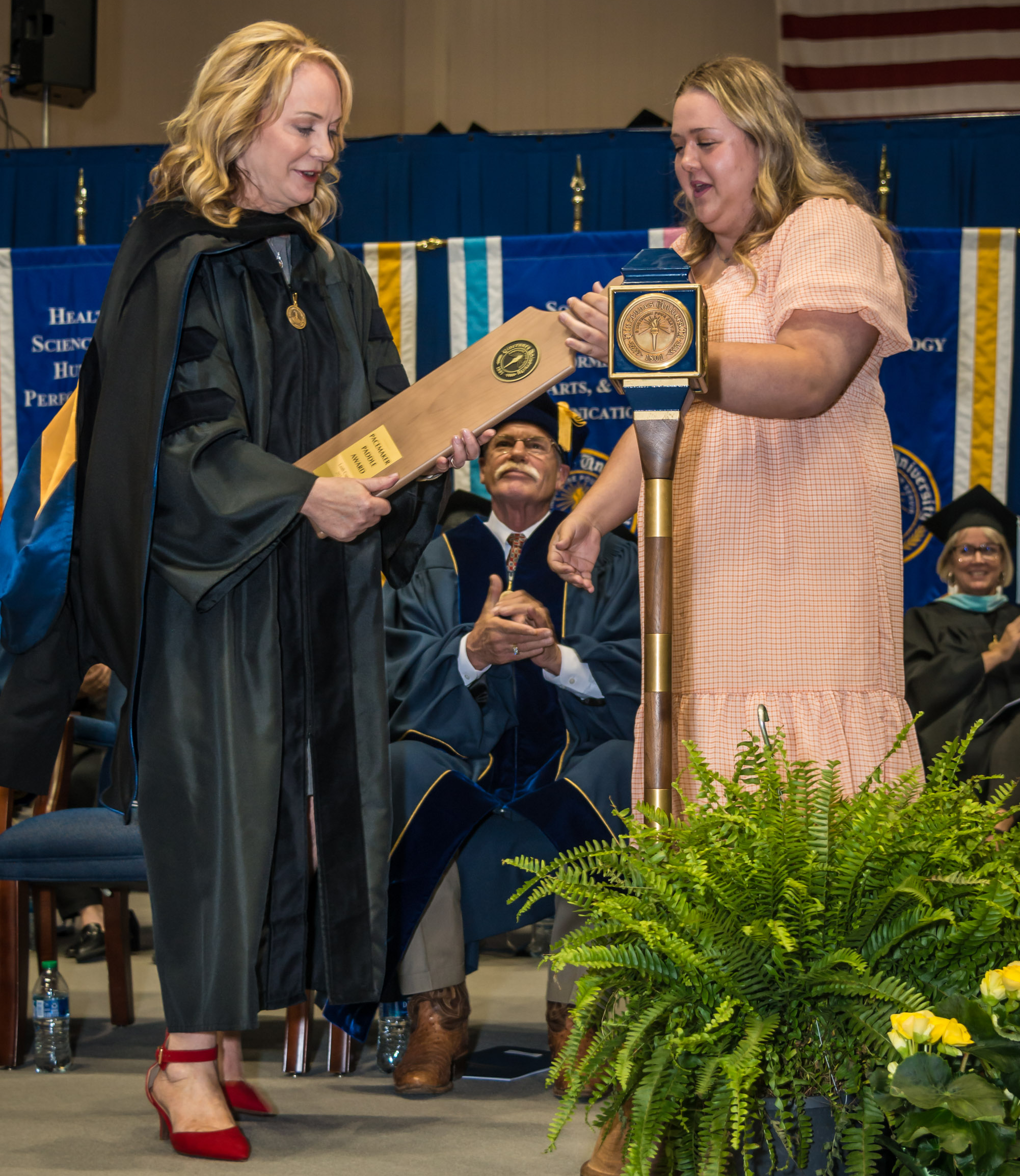 A VU student hands Leah Curry who is dressed in a doctorate robe and hood a paddle award.